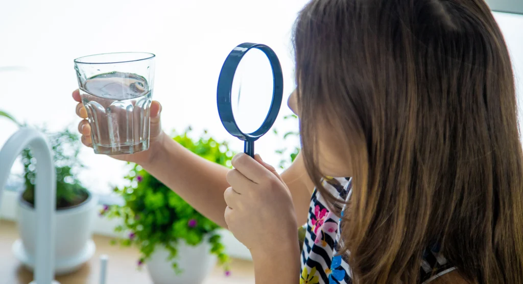 Child examining a glass of well water with a magnifying glass — coliform and E. coli cannot be seen