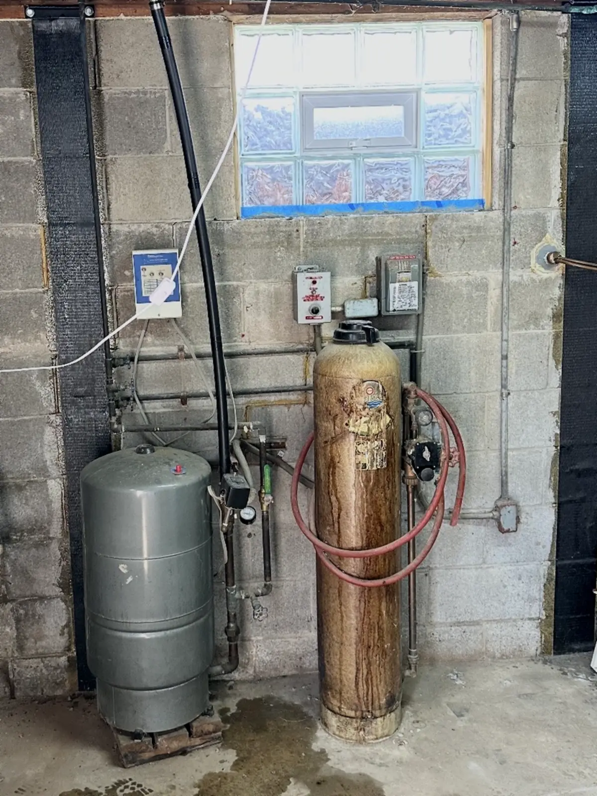 Older whole-house water treatment tank and well pressure tank on a block basement wall in Southwestern Pennsylvania, a system still sound enough that a repair is the right call