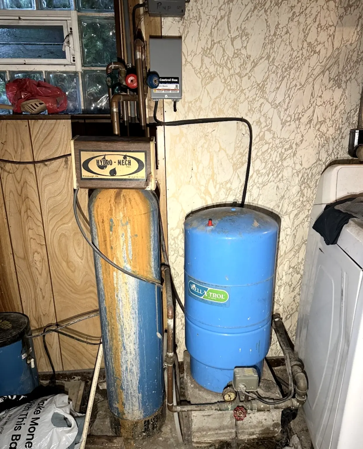 Heavily rust-stained whole-house water treatment tank next to a well pressure tank in a Southwestern Pennsylvania basement, a system far past the point where a repair is worth doing